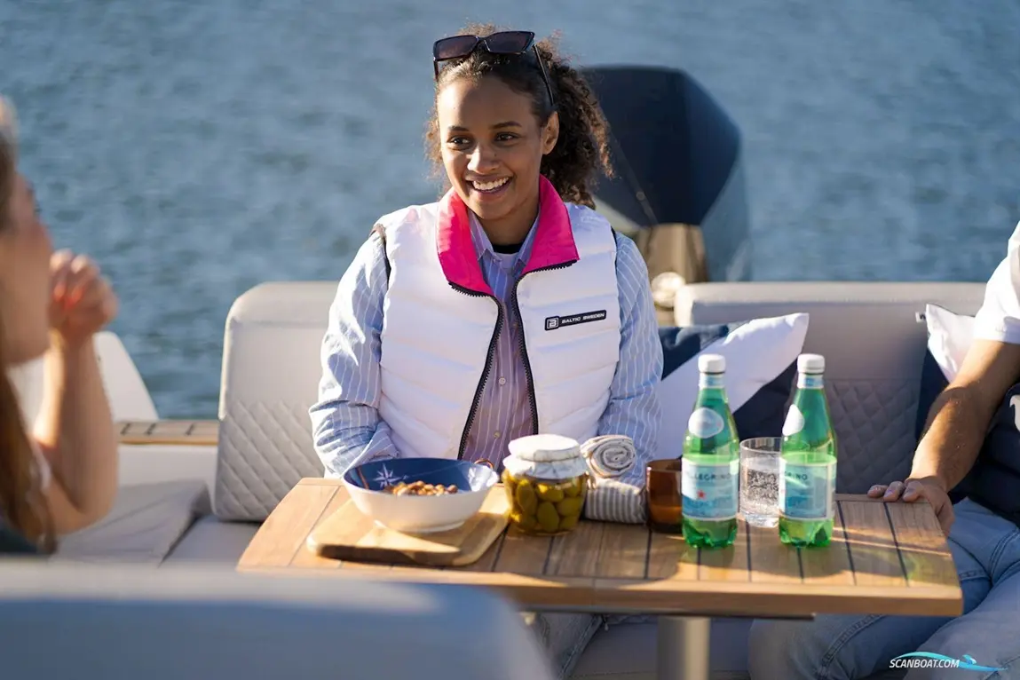 A woman in a white vest smiles at a table with snacks, drinks, and a scenic background on a boat.